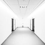 A minimalist corridor featuring two elevators on either side, white walls, and smooth flooring, illuminated by bright ceiling lights, creating an open and modern atmosphere.