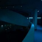 A dimly lit modern interior featuring a spiral staircase, smooth white walls, and subtle blue lighting, creating a serene and minimalist atmosphere.