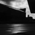 A monochrome photograph of modern architectural buildings with angular shapes, set against a cloudy sky. The foreground features wet pavement with reflective patches of light.
