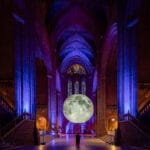 A large, illuminated replica of the moon is displayed in the interior of a grand, stone cathedral, surrounded by purple and blue lighting, with a person standing in front of it looking up, and stained glass windows visible in the background.