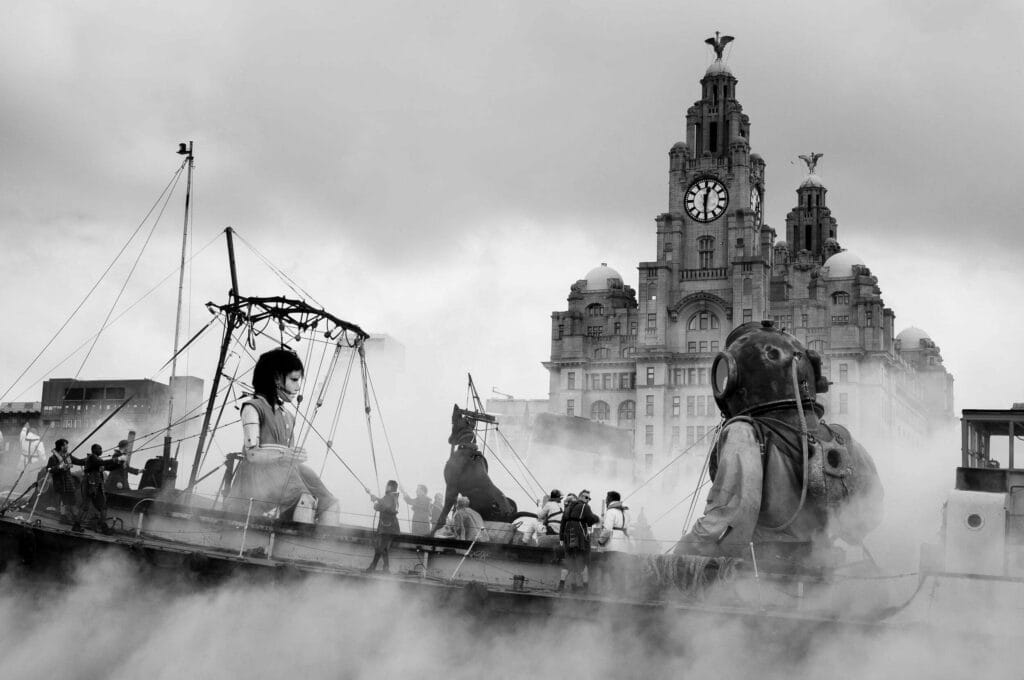 A large puppet of a girl and a man in a vintage diving suit on a boat, surrounded by fog, with the Royal Liver Building in the background, set against a cloudy sky in monochrome.