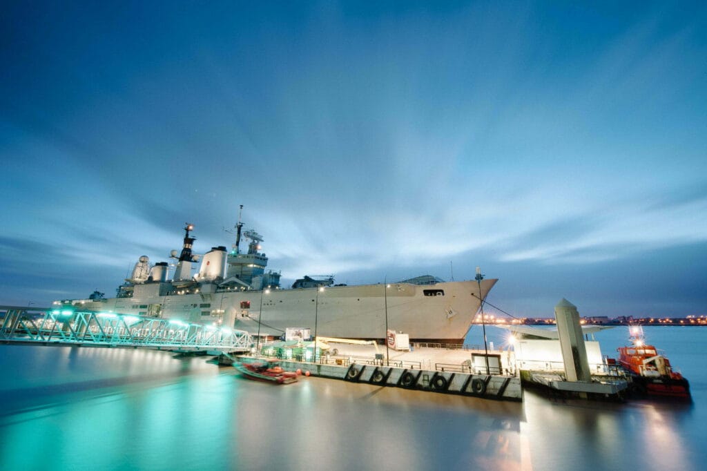A Royal Navy aircraft carrier docked in Liverpool, with illuminated surroundings reflecting on the water, and a small boat near the quay in the foreground under a dusk sky.