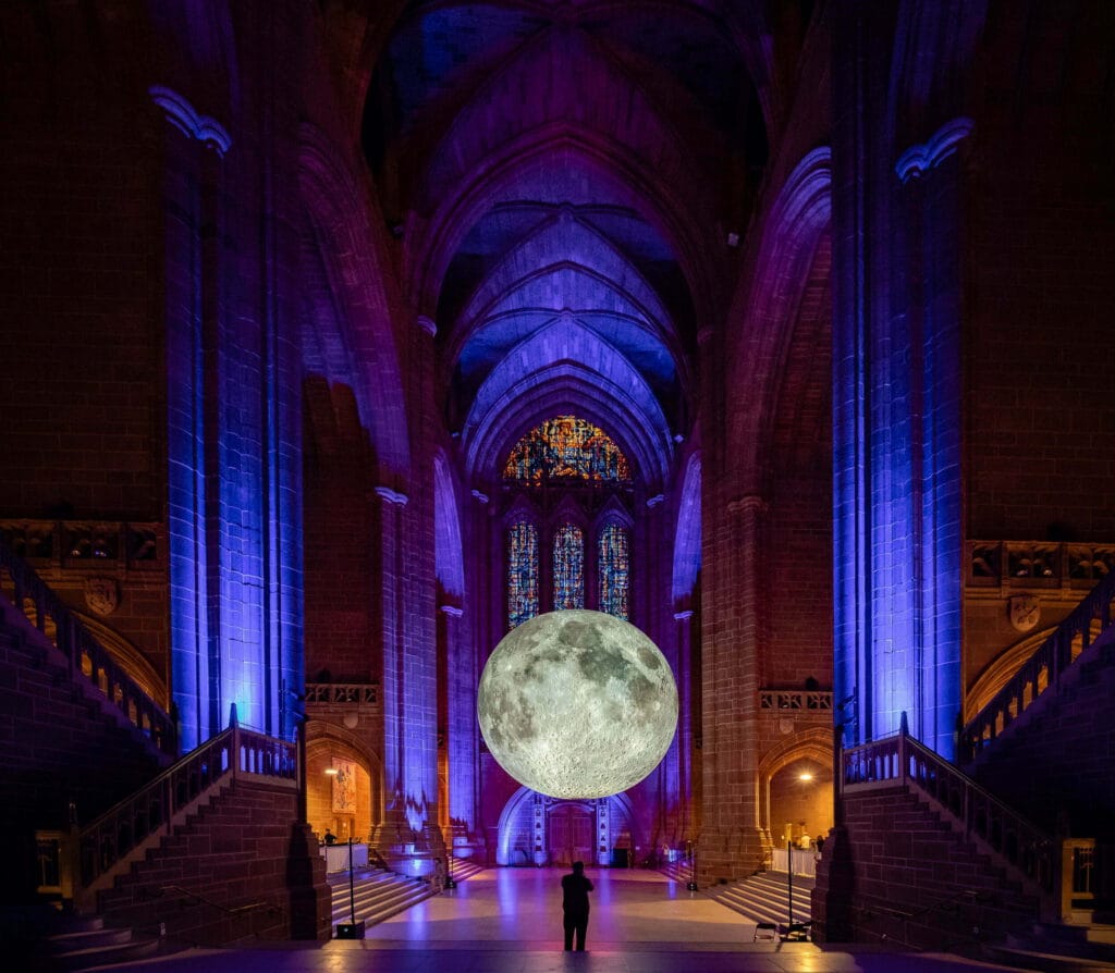 A large illuminated replica of the moon suspended in a Gothic-style cathedral, highlighting the intricate stained glass windows and atmospheric purple and blue lighting, with a silhouette of a person standing below.