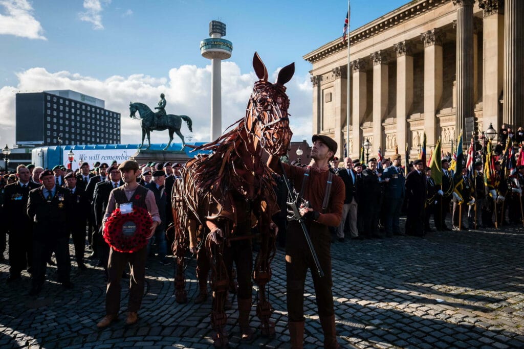 A crowd gathers in Liverpool for a commemorative event, featuring a large bronze horse sculpture, a man holding a poppy wreath, and a smaller horse figure made from metal. Flags and uniforms suggest a military theme, with the Royal Liver Building and Radio City Tower visible in the background.