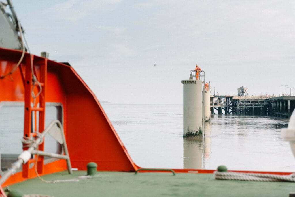View from a boat showing the Liverpool cruise terminal with a concrete pier and a person in an orange uniform standing on the pier, surrounded by calm water under a light sky.