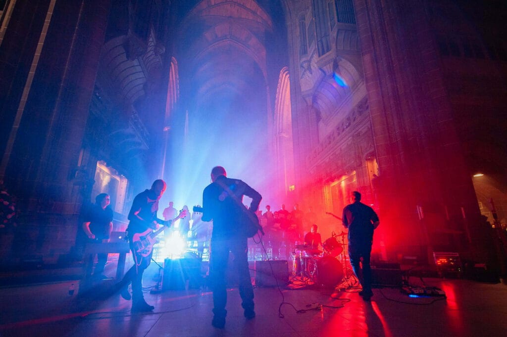 A live music event inside a large, atmospheric cathedral, featuring musicians on stage illuminated by dramatic blue and red lighting, with a choir visible in the background and smoke effects enhancing the mood.