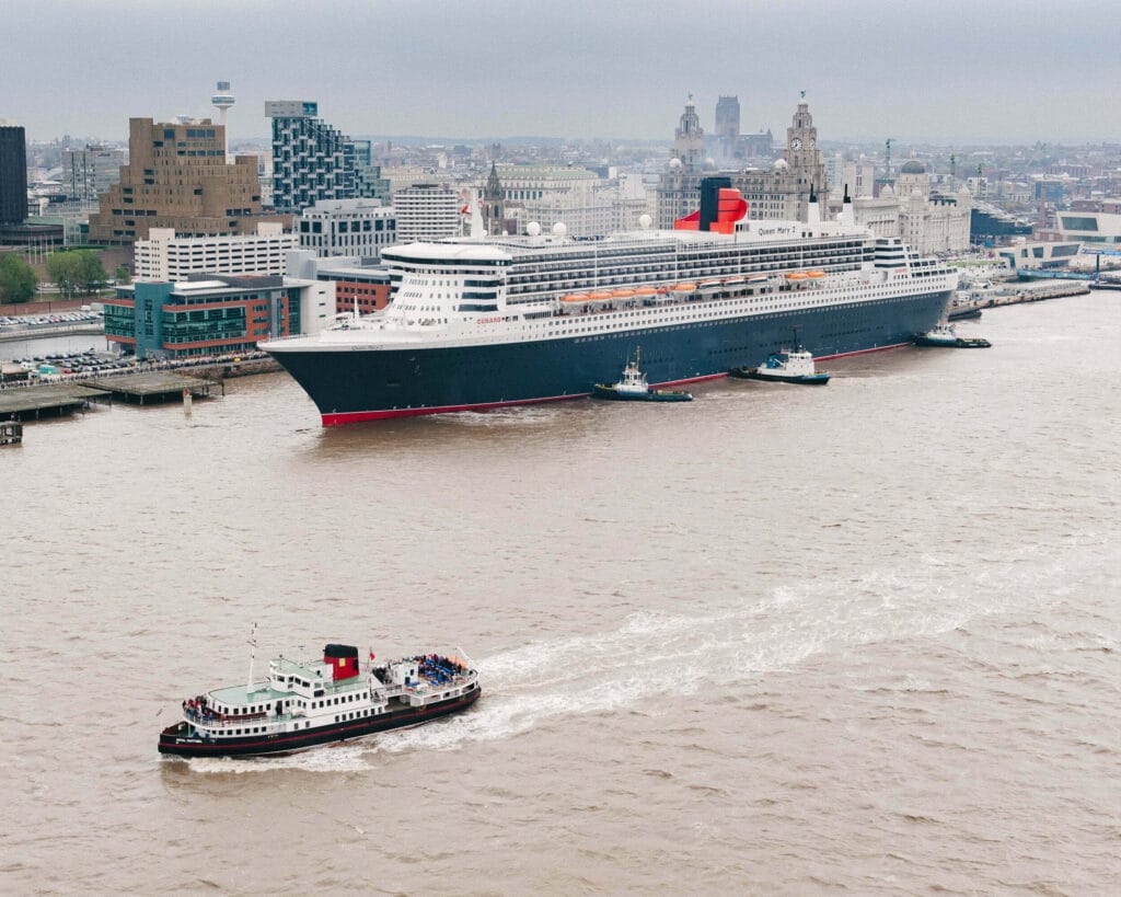 The Cunard Line's Queen Mary 2, a large ocean liner, is seen navigating the Mersey River in Liverpool, flanked by several tugboats, with the city's skyline in the background and a smaller boat in the foreground carrying passengers.