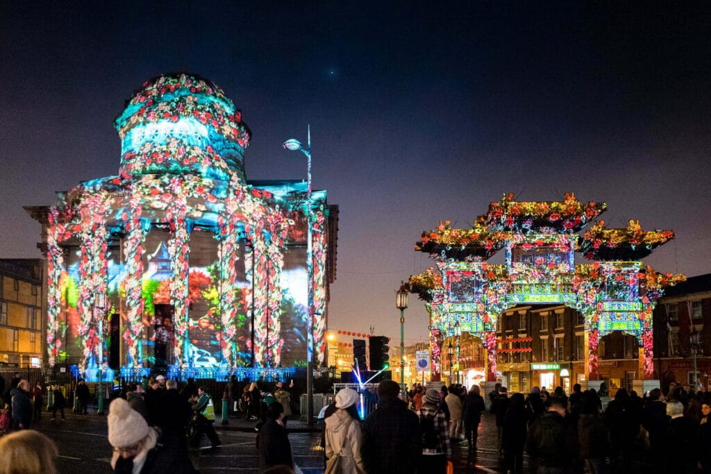 A vibrant evening scene from a Liverpool event, featuring a historic building adorned with colorful floral projections on the left, and an ornate illuminated archway on the right, surrounded by a crowd of people enjoying the festivities.