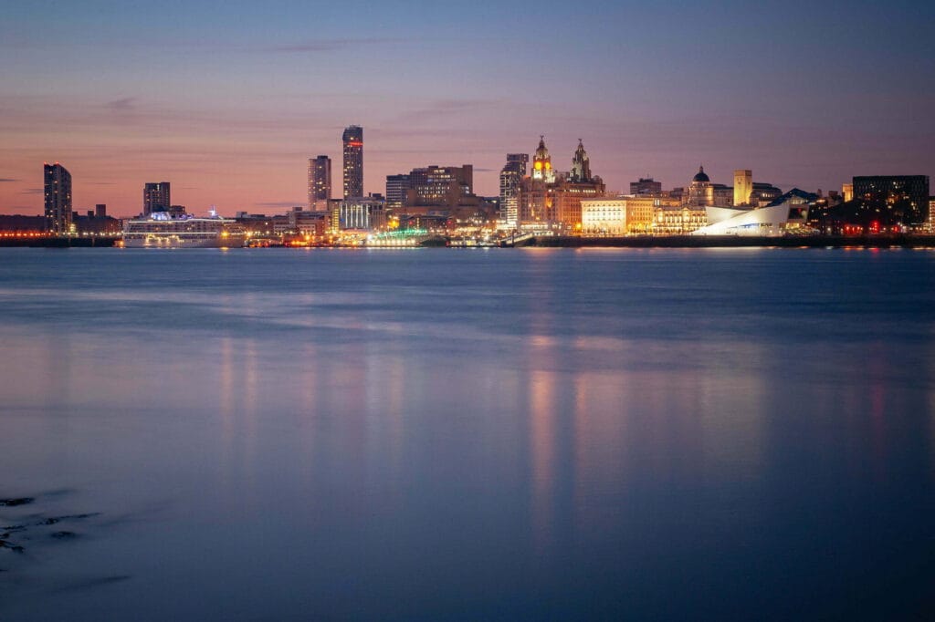 A twilight view of Liverpool's skyline featuring illuminated buildings along the waterfront, with reflections in the water and a prominent, modern structure on the right side.