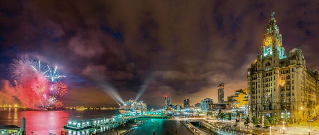 The cruise ship Celebrity Infinity is docked in Liverpool, illuminated against a night sky filled with fireworks, with the iconic Royal Liver Building prominently featured on the right.