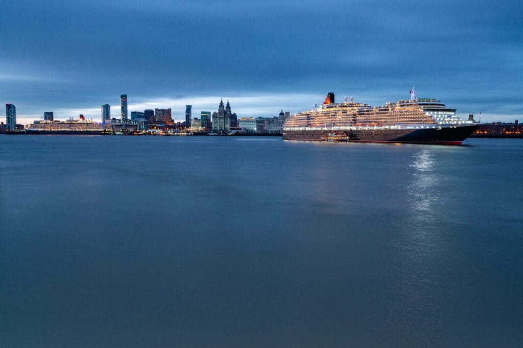 Two illuminated Cunard cruise ships docked in the Mersey River near the Liverpool skyline during twilight, showcasing a vibrant cityscape in the background.