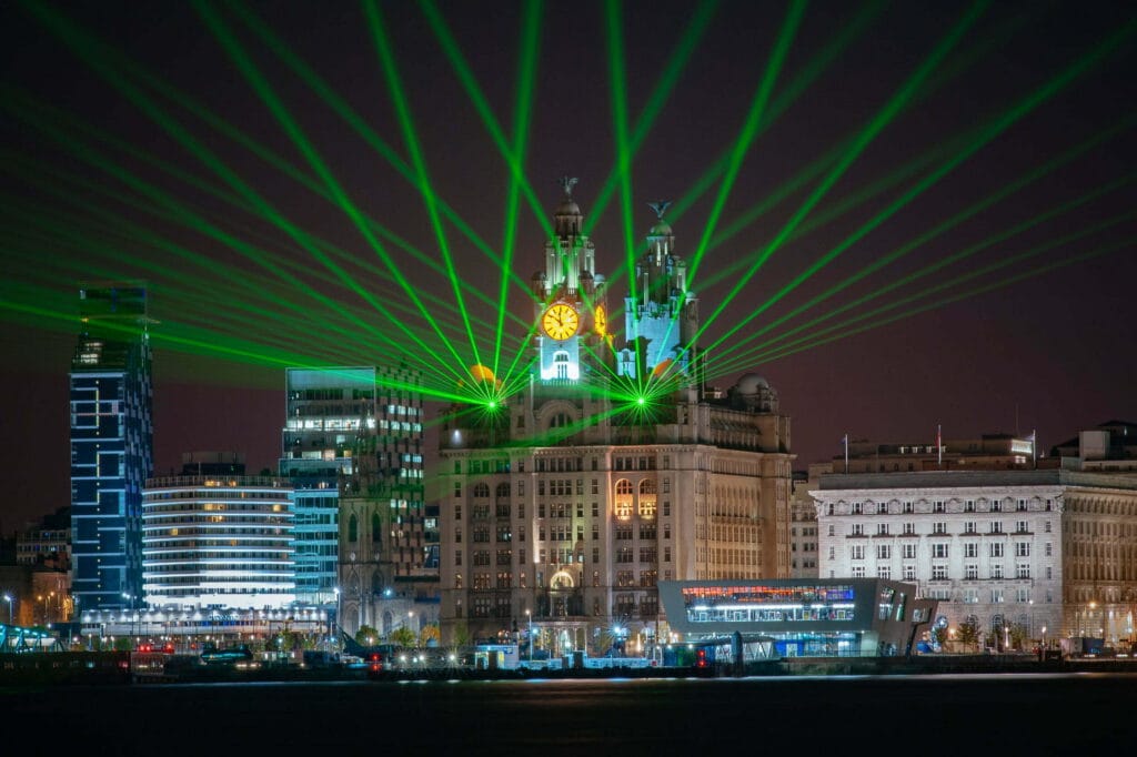 A nighttime view of Liverpool's waterfront featuring the iconic Royal Liver Building illuminated by green laser beams, surrounded by modern skyscrapers and the waterfront promenade.