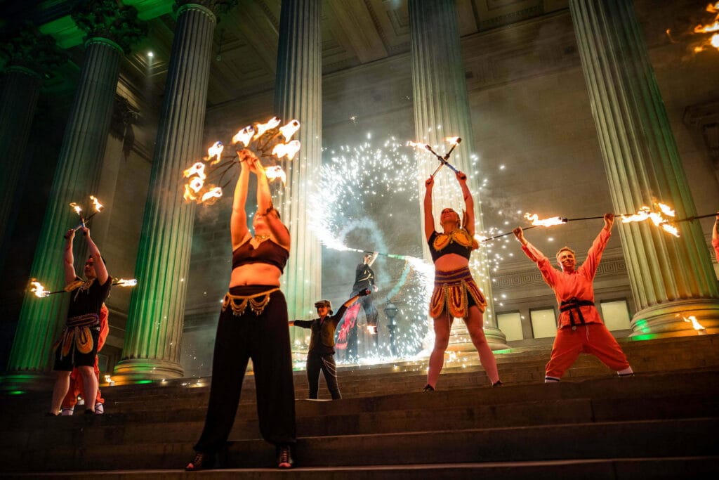 Performers at a Liverpool event showcase a fire dance in front of grand columns, with some holding flaming staffs aloft and others creating sparks in the background, all dressed in vibrant costumes.