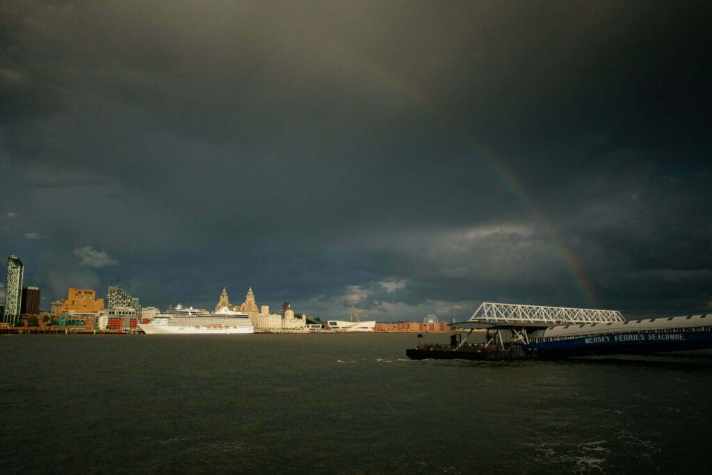 A cruise ship docked in Liverpool, surrounded by modern and historic buildings under a dramatic sky with dark clouds and a rainbow in the background.