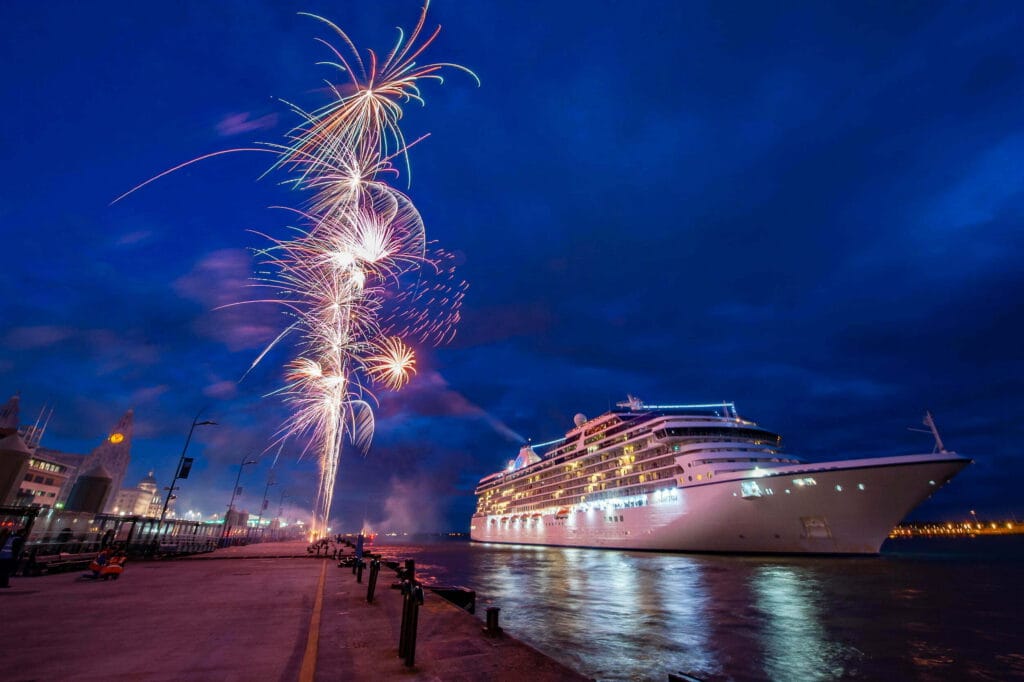 A cruise ship illuminated at night, docked in Liverpool, with colorful fireworks bursting into the sky above, set against a dark blue backdrop.