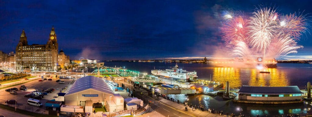 A vibrant cityscape of Liverpool at night featuring an ACL cargo ship docked with fireworks illuminating the sky. The iconic Royal Liver Building is visible in the background alongside the Liverpool Cruise Terminal, as crowds gather along the waterfront.