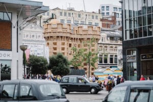 A bustling street scene in Liverpool featuring a large, decorative wooden structure resembling a castle, surrounded by greenery and onlookers, with a mixture of modern and historic buildings in the background, including a sign that reads 'Beautiful World Where are You?' and black taxis driving in the foreground.