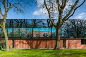 Exterior view of the Whitworth Gallery in Manchester, showcasing a modern glass facade reflecting nearby trees and a blue sky, with a contrasting brick lower section and landscaped greenery in the foreground.