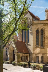 A view of a historic building at the University of Manchester, featuring intricate stonework, stained glass windows, and a red-tiled roof, surrounded by lush green trees and planters with flowers.