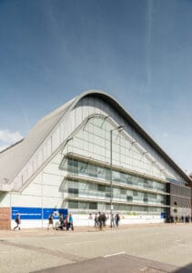 A modern swimming baths building on Oxford Road in Manchester, featuring a distinctive curved roof and large glass windows, with pedestrians and cyclists in front on a sunny day.