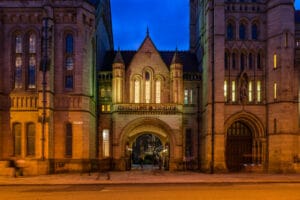 The grand entrance of the University of Manchester, featuring an archway with the name prominently displayed, flanked by ornate stone towers and illuminated windows, set against a twilight blue sky.