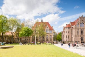 A sunny view of the University of Manchester campus, featuring historic brick buildings with red roofs, surrounded by lush green trees and grass, with people walking and cycling along pathways.
