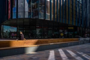 A person sits on a wooden bench outside the HOME gallery in Manchester, with large glass doors displaying promotional posters and a reflection of the surrounding buildings, and contrasting shadows on the pavement.