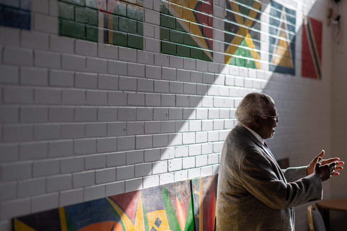 Dr Julius Garvey standing next to a white brick wall as a beam of light shines across it at an angle. There are flags on the wall.