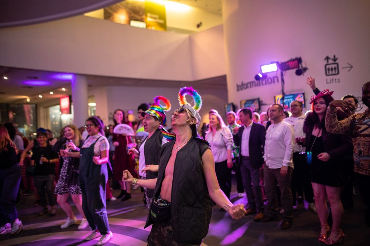 People dancing at a live music gig inside the Museum of Liverpool. Two people, centre frame, are wearing rainbow angel halos.