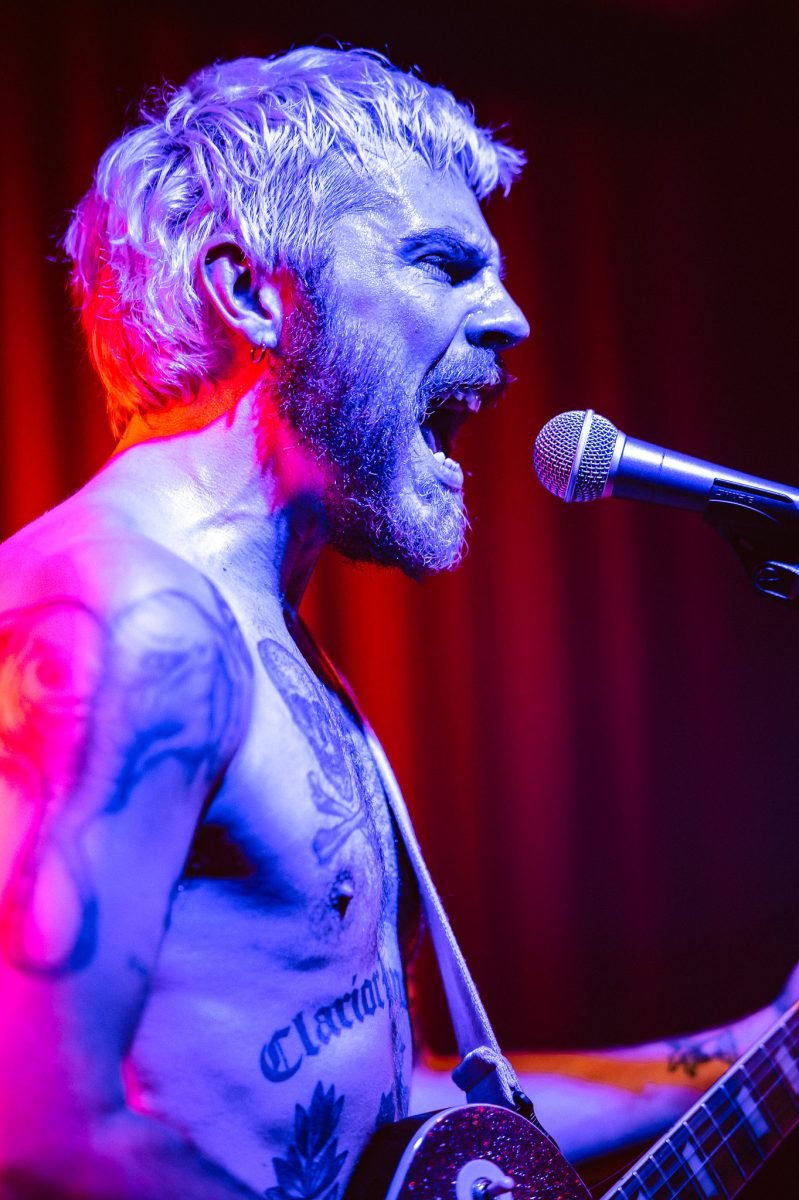 A punk singer singing while lit in blue light in front of a red curtain.