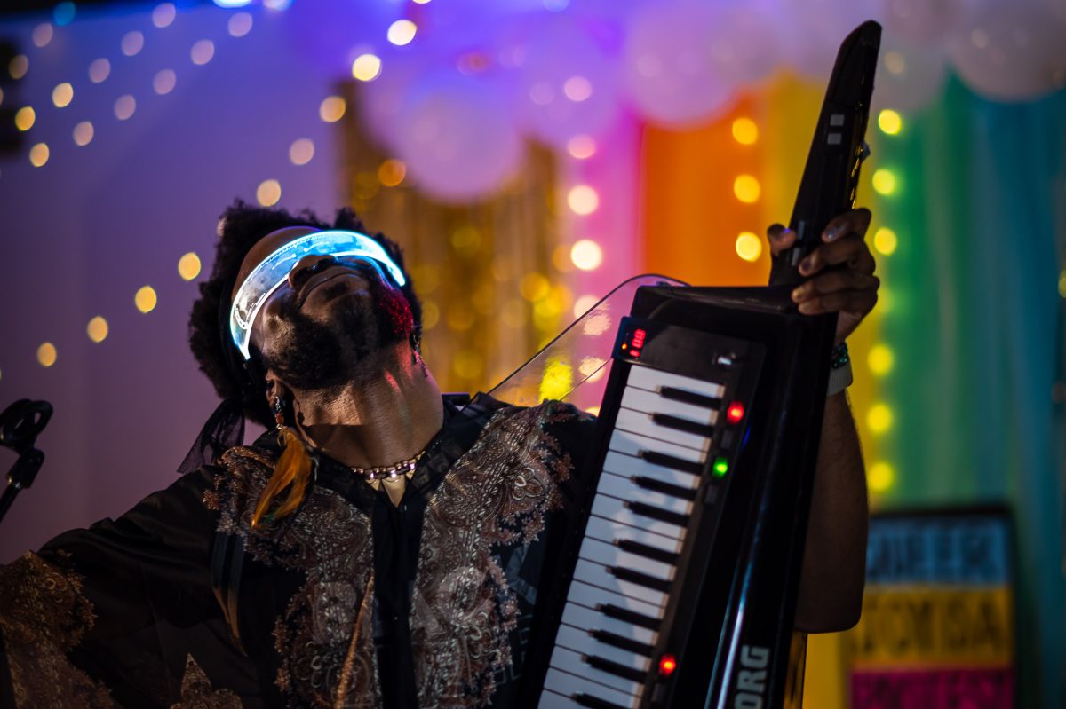 A black person playing a keytar while wearing futuristic illuminated eye wear in front of a rainbow flag.