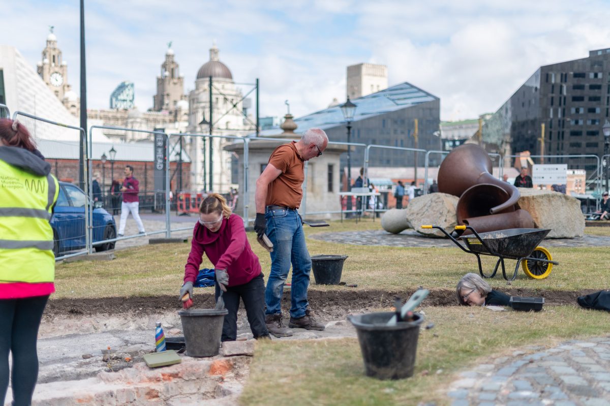 People doing archaeology on a green space in the Royal Albert Dock. Some are wearing hi-vis jackets. Others are digging with buckets and spades. In the distance is the Liverpool waterfront.