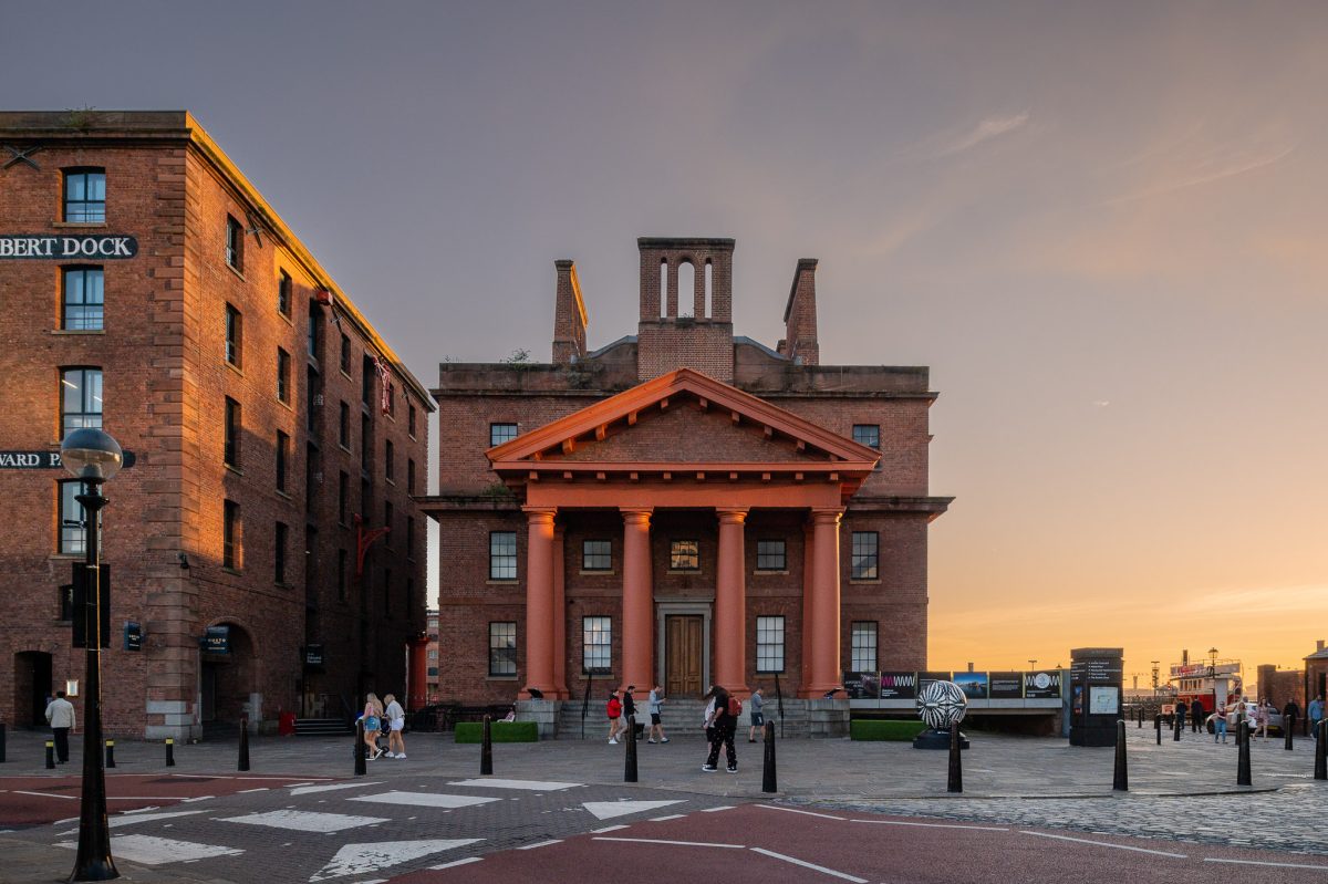 Sunset outside the Martin Luther King Building at Royal Albert Dock. The sun sets to the right and there is an orange glow in the sky. The building has 4 columns at the front and a distinctive style.