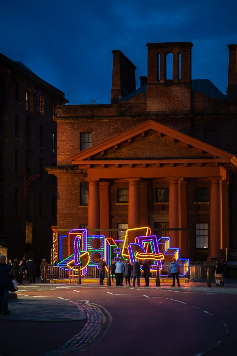 Lighting installation comprising of various colourful shapes lit up at dusk outside the Martin Luther King building. There are people enjoying it.