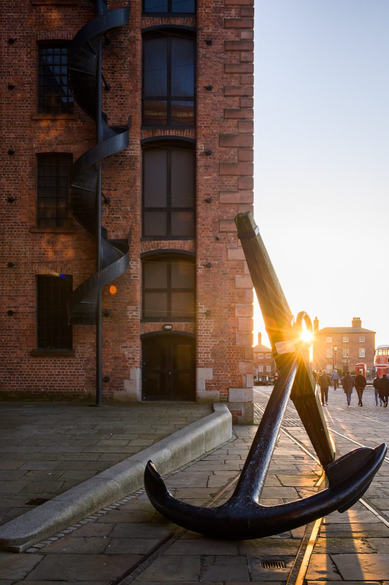 Looking at the side of the Maritime Museum as the sun sets through the top of the large anchor resting outside it. 