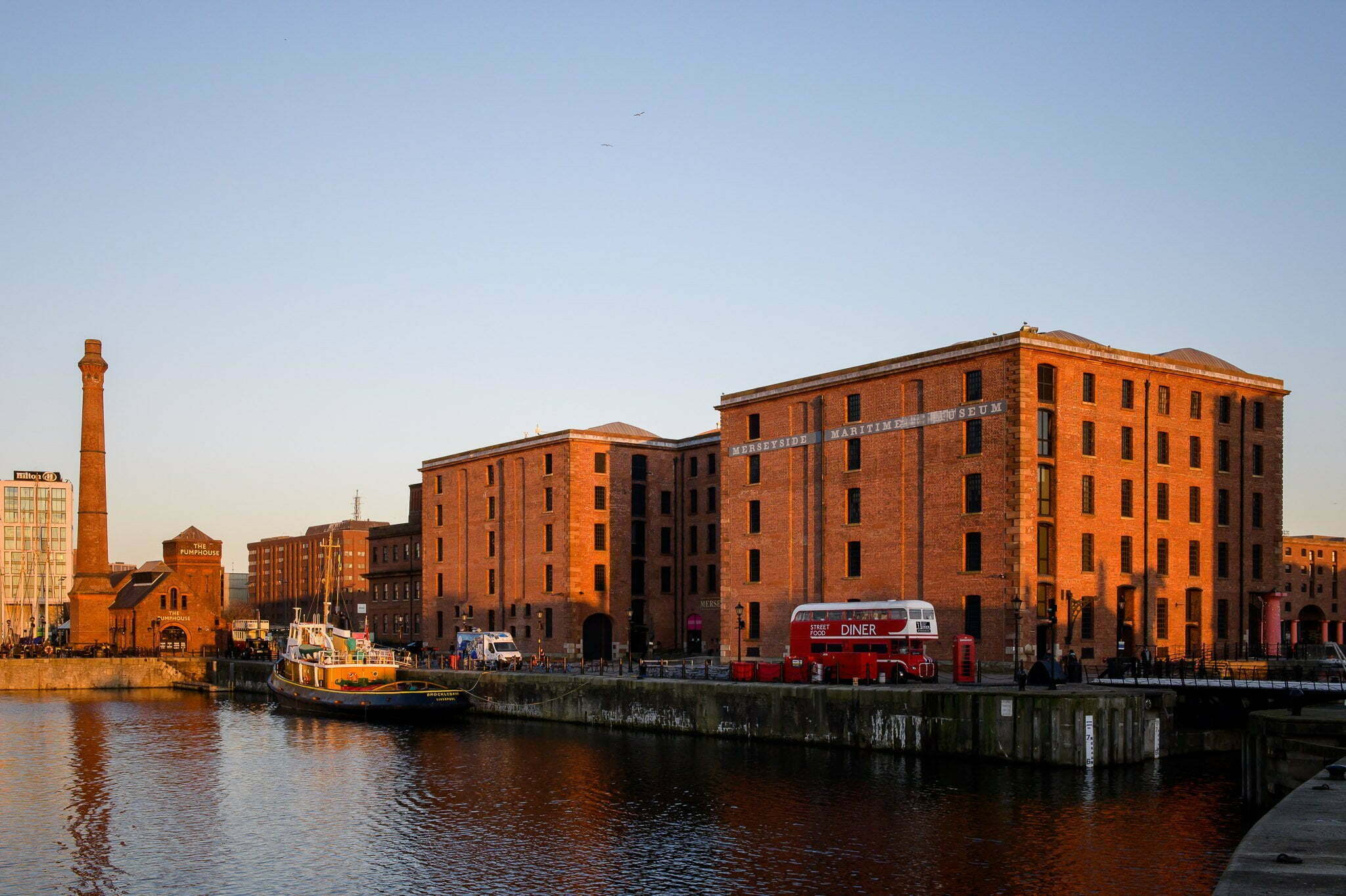 As the sun sets the red brick Maritime Museum glows orange against a blue sky. The dock water is calm.