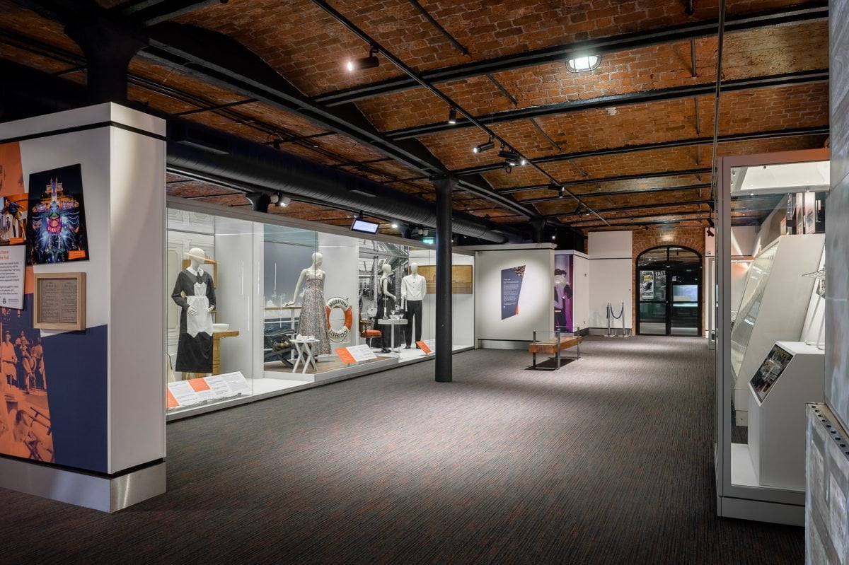 Inside a museum gallery. It has a red brick ceiling. There are mannequins inside the display window wearing various clothes that you might find on a cruise liner in the early 1900s.

