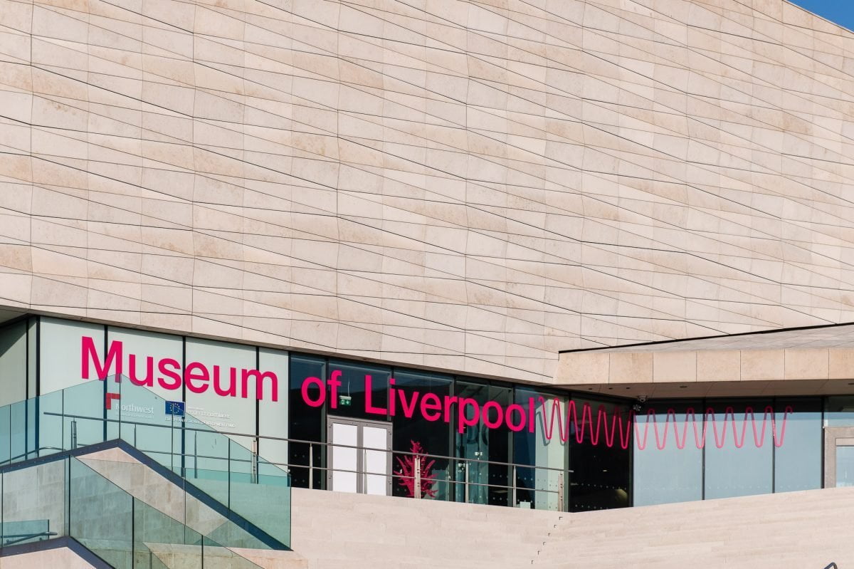 Standing on the steps of the Museum of Liverpool. There is a sign in the window saying "Museum of Liverpool" in purple / pink. 