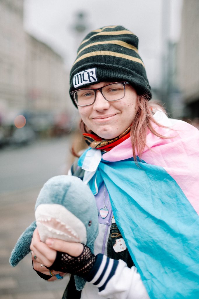 A person in a trans flag and MCR beanie hat looks to the camera and holds up an Ikea plush shark. 
