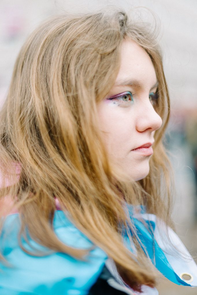A side profile of a young female presenting person, with blonde hair. They have pink and blue trans flag colour eye shadow on and a trans flag around them.
