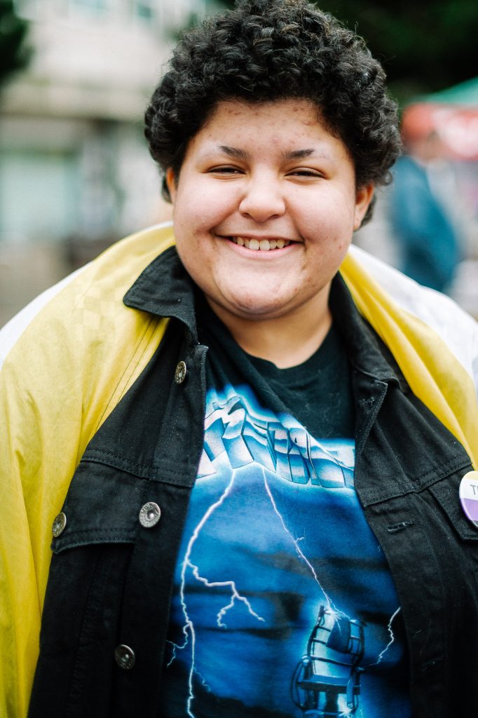 A person is draped in a non-binary flag and smiles to the camera.
