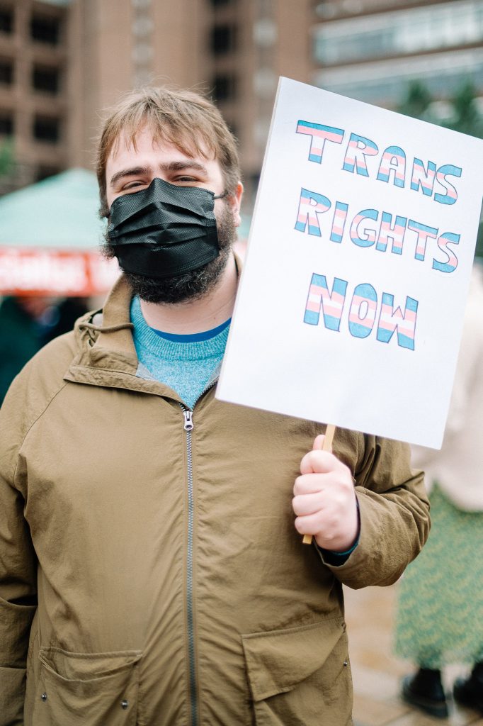 A male presenting white person in a mask holds up a sign saying "Trans rights now."
