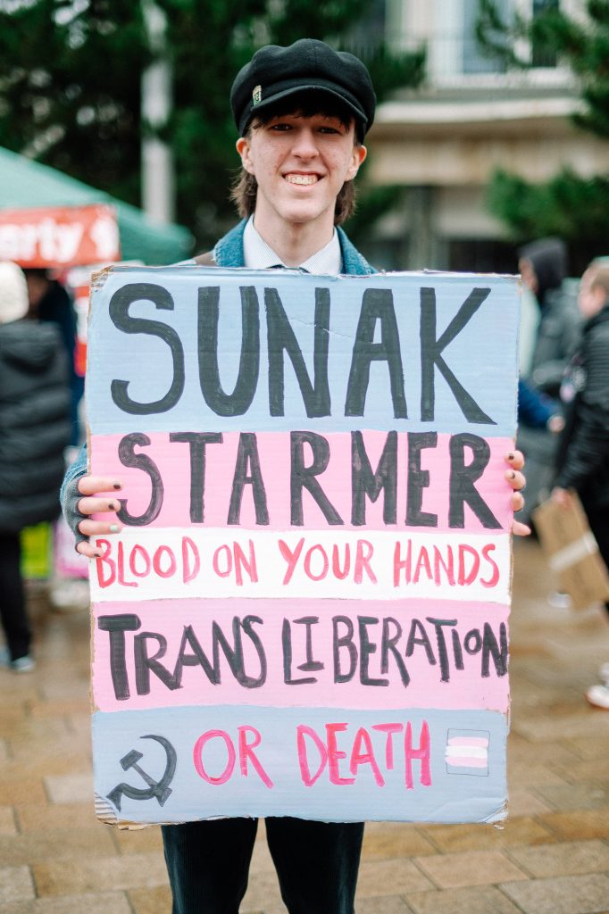 A 30 something male presenting person wearing a cap holds up a sign saying "Sunk. Starter. Blood on your hands. Trans liberation or death."
