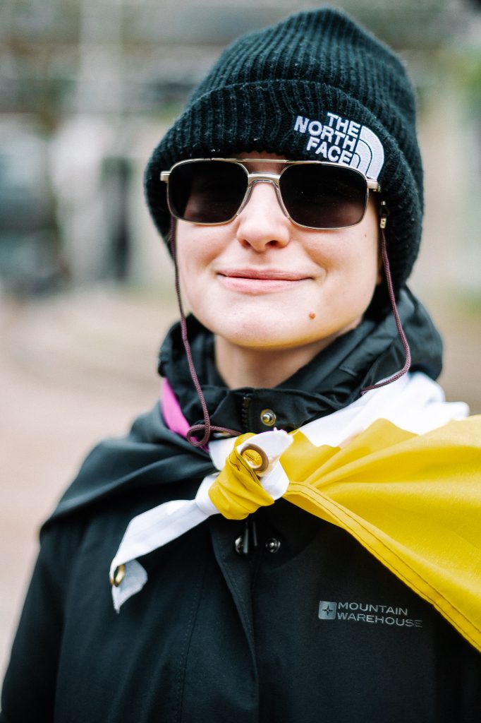 A 20 something white person smiles to camera. They are draped in a non-binary flag wearing all black clothes and a beanie hat.
