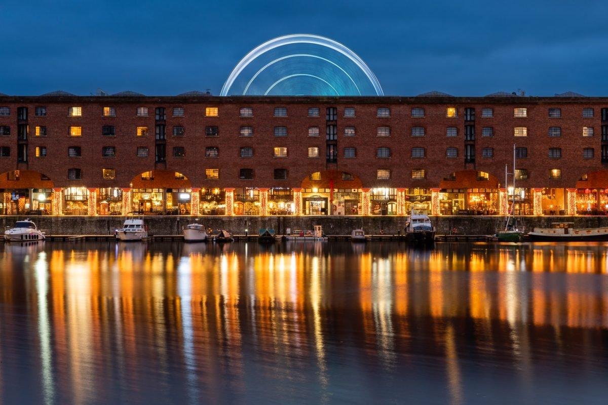 Looking across the Albert Dock towards the restaurants at dusk. The water reflects the lights from the lit up colonnades and stores. There is a large wheel spinning behind the buildings.