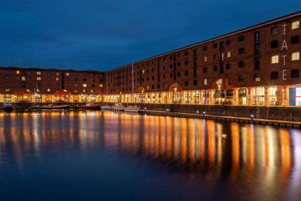 Looking across the Albert Dock towards the restaurants at dusk. The water reflects the lights from the lit up colonnades and stores. There is a large wheel spinning behind the buildings.