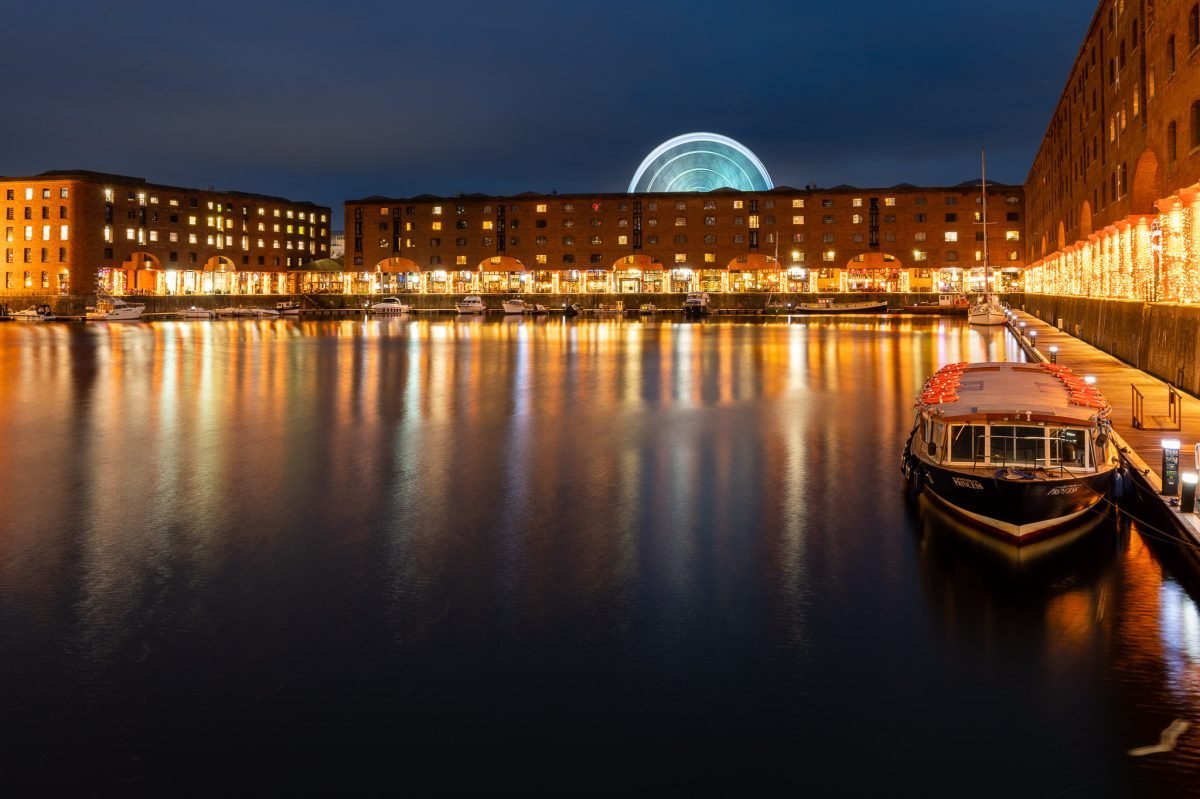 Wide view across the Albert Dock towards the restaurants at dusk. The water reflects the lights from the lit up colonnades and stores. There is a large wheel spinning behind the buildings.