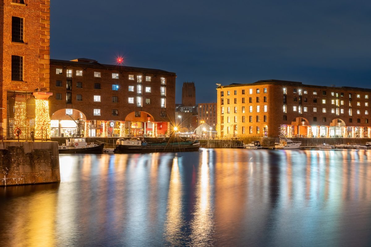 Looking across the Albert Dock at dusk. There are old dock warehouses repurposed as restaurants on either side. In the distance is Liverpool Anglican Cathedral.