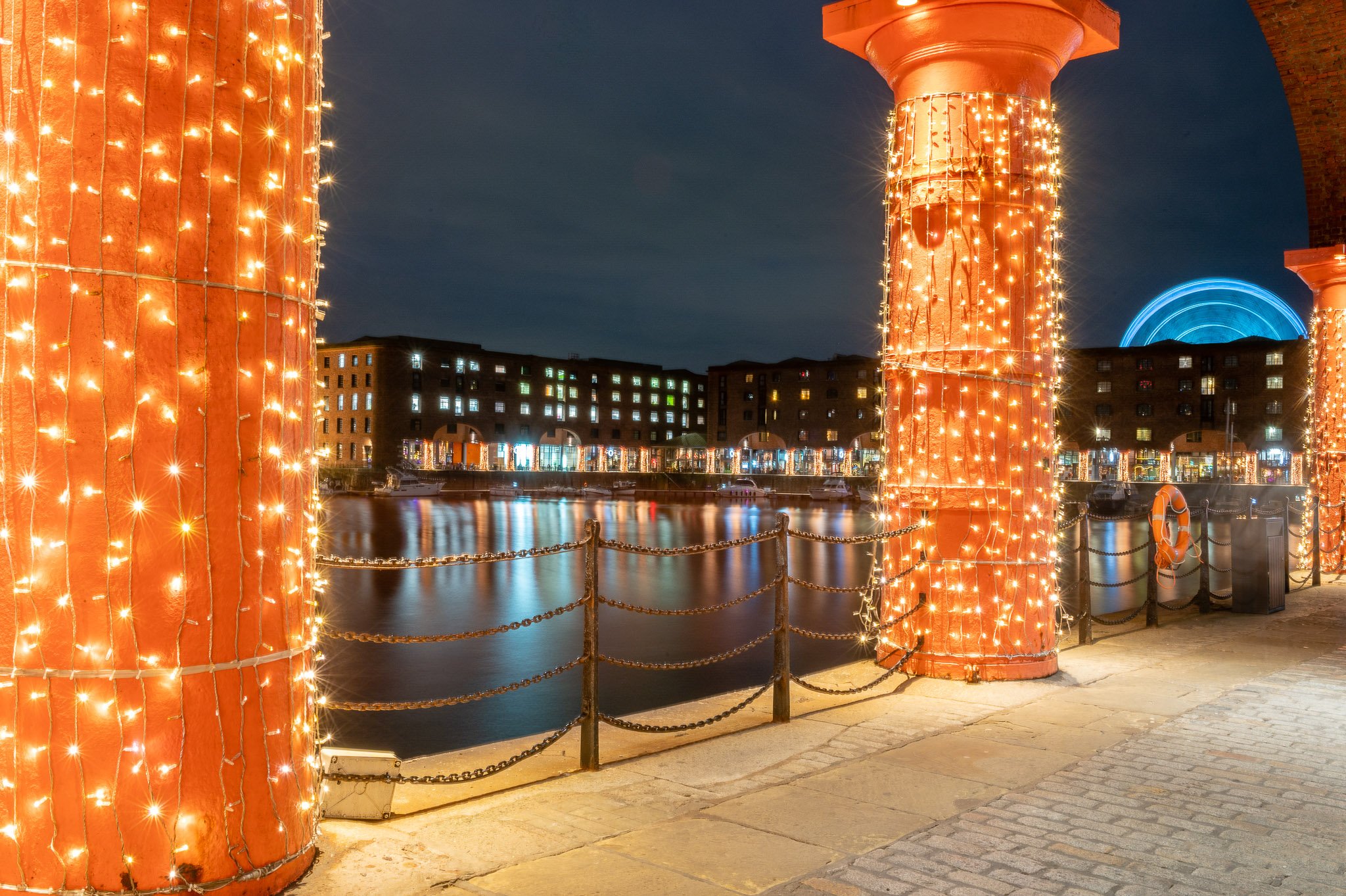 Dusk view of Albert Dock from the colonnades area. Behind the buildings, a viewing wheel spins. There are fairy lights on each column of the colonnades.