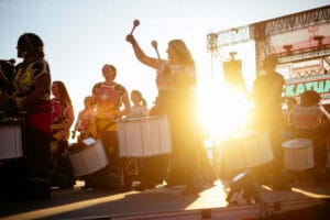 A group of women in vibrant outfits play drums during a performance at sunset, with sunlight creating a glowing effect around them.